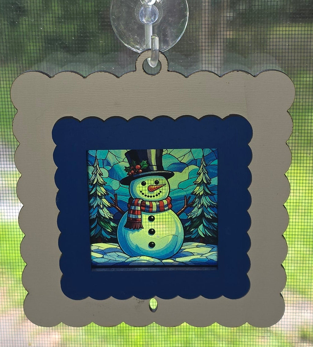 Laser-cut wood interchangeable Christmas suncatcher frame 4.5" with snowman insert, white scalloped frame and blue inner mat, hanging in window with suction cup – Illuminated Engraving

